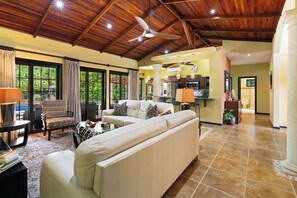 A spacious living area with a high ceiling and a ceiling fan, a large sofa, a dining table, and a kitchen in the background.