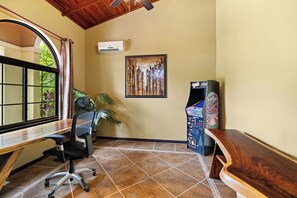 Una habitación con un escritorio de madera, una silla, un ventanal grande, un equipo de aire acondicionado, una obra de arte enmarcada y una máquina expendedora.