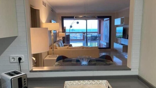 A modern hotel room with a large window, a sofa, a dining table, and a TV.