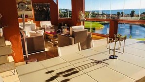 A spacious room with a large window overlooking a pool and greenery. There is a dining area with a table and chairs, and a seating area with sofas.