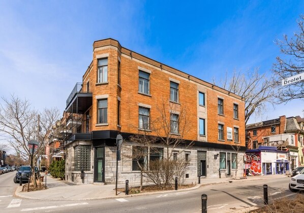 A multi-story building with a brick facade, a visible ground-floor entrance, and a street view with parked cars and a clear sky.