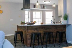 Une cuisine dotée d’un îlot central en bois, de tabourets de bar, de luminaires suspendus et d’un pare-feu sur lequel est inscrit le mot « CUISINE ».