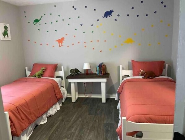 A room with two beds, each with a red bedspread, a small table with a lamp and books, and a wall decorated with animal stickers.