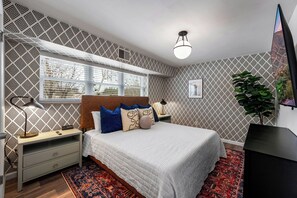 Ein Schlafzimmer mit gemustertem Wandpapier, einem Bett mit vielen Kissen, einem Nachttisch mit Lampe, einem Fenster mit Jalousien und einer Pflanze.