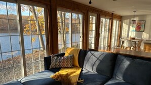 A cozy living room with a large window overlooking a lake, a dark blue sofa, and a dining area with a table and chairs. - Lakefront Connecticut Oasis - Updates throughout  (Oxford)