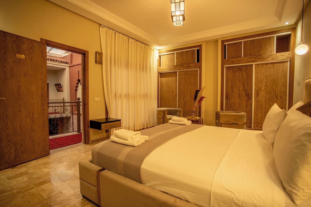A hotel room with a large bed, a wooden wardrobe, a small table, and a chair.