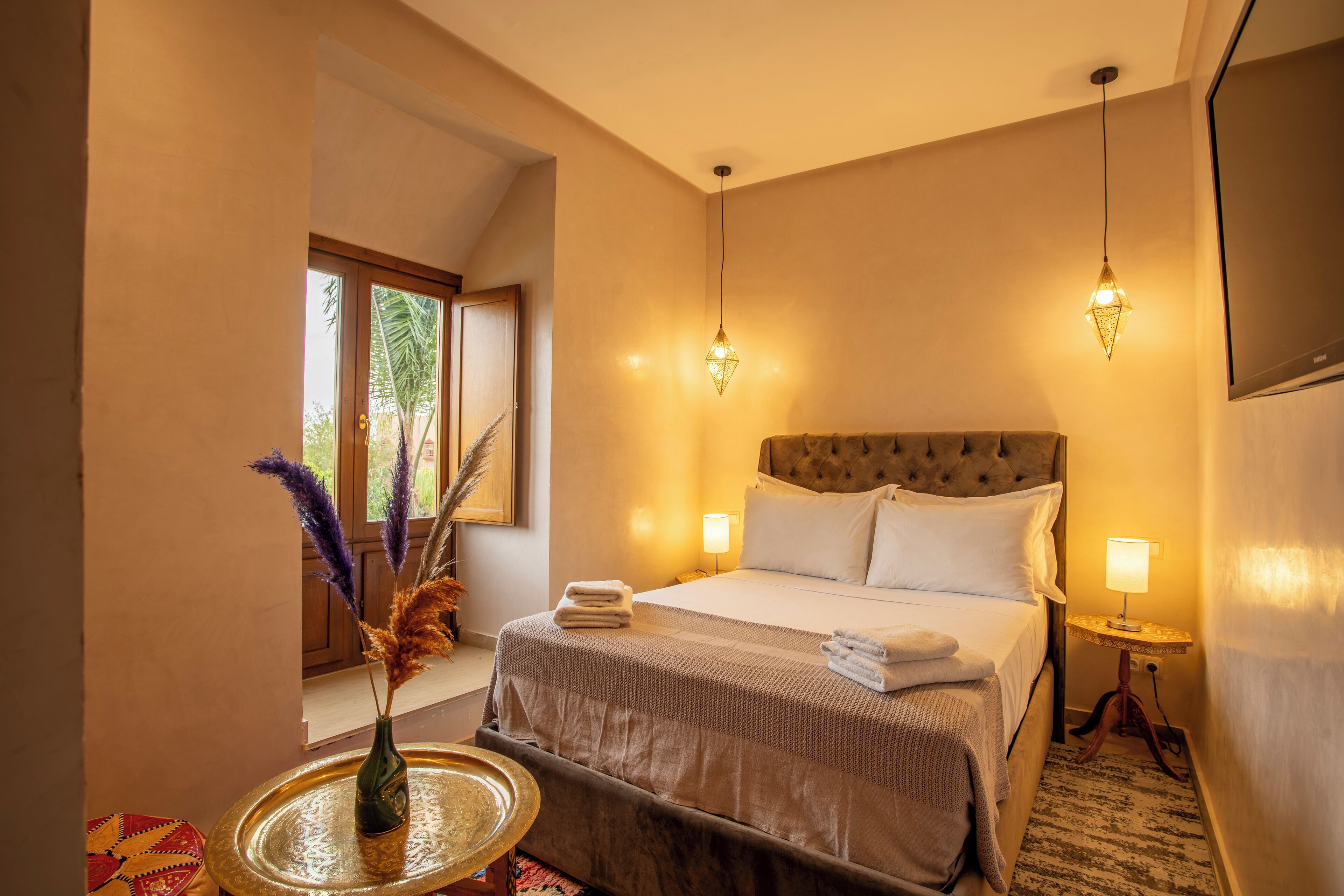 A hotel room with a large bed, bedside lamps, a small table with a vase, and a window with a view of greenery.