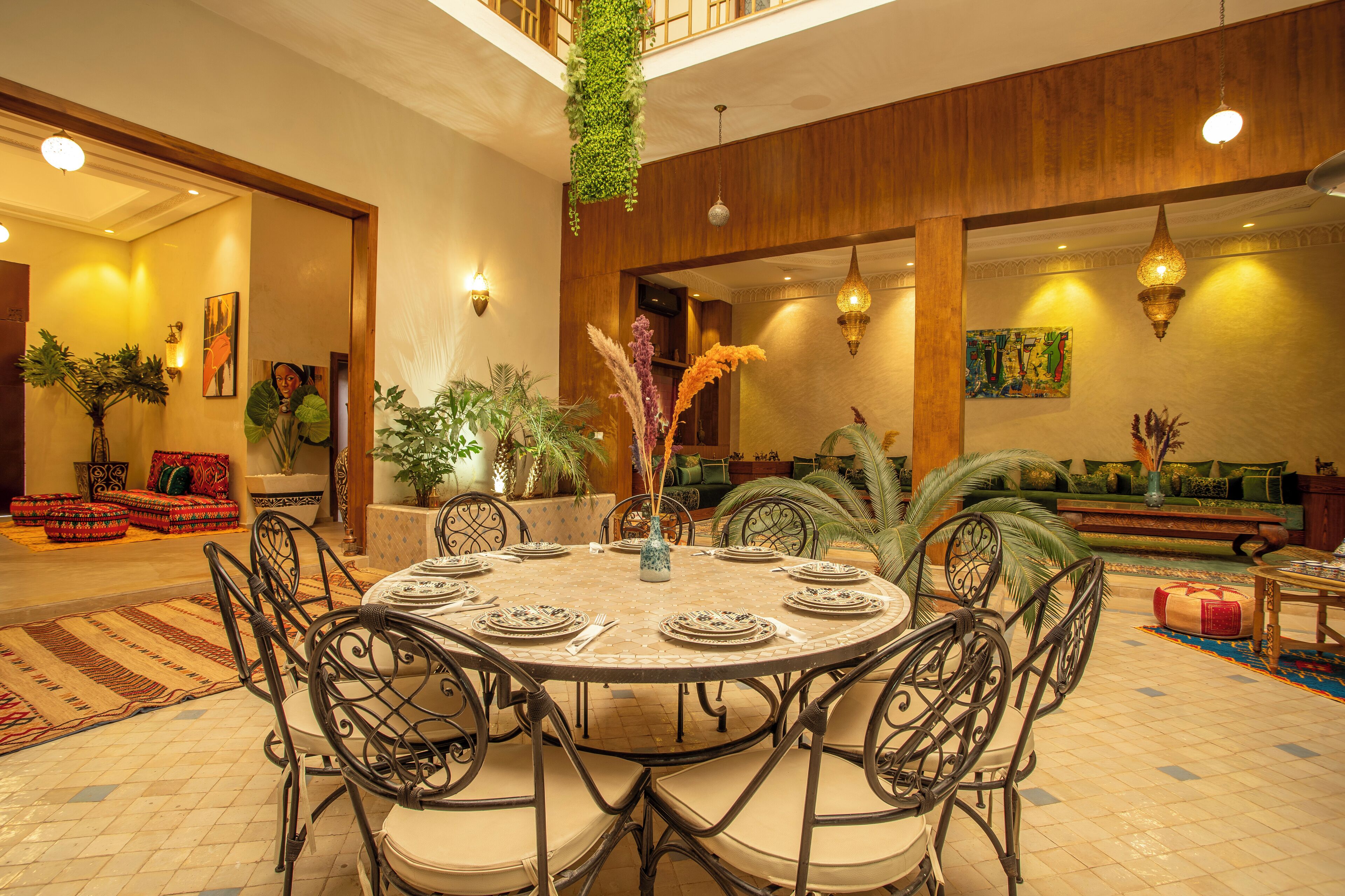A dining area with a round table, chairs, and a vase with flowers. There are plants and a sofa in the background.