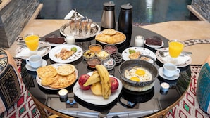 A breakfast table with various dishes, including pancakes, eggs, and fruits, set on a terrace with a pool in the background.