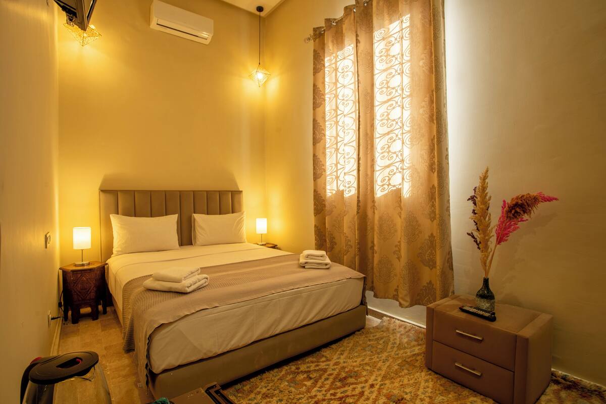 A hotel room with a bed, bedside tables, a nightstand, a dresser, a window with curtains, and a vase with flowers.