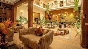 A spacious lobby with a high ceiling, a central seating area with a sofa and armchairs, a dining area with tables and chairs, and a balcony with a railing.