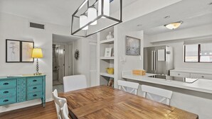 A dining area with a wooden table and white chairs, a turquoise cabinet with drawers, and a white countertop with a sink. - Sea La Vie: 2Bed Gulf-Adjacent Condo - by SkyRun Anna Maria (Bradenton Beach)