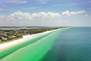 A coastal view with a beach, clear turquoise waters, and a town in the background. - Sea La Vie: 2Bed Gulf-Adjacent Condo - by SkyRun Anna Maria (Bradenton Beach)