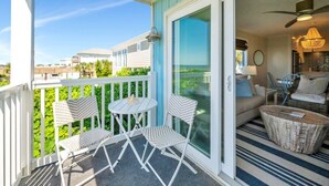 A balcony with white chairs and a table, a sliding glass door leading to a living area with a sofa, a lamp, and a ceiling fan. - Oceanfront in Ocean Isle Beach! "C2 Shining Sea" - 3 bed 2 bath Condo - Sleeps 8 (Ocean Isle Beach)