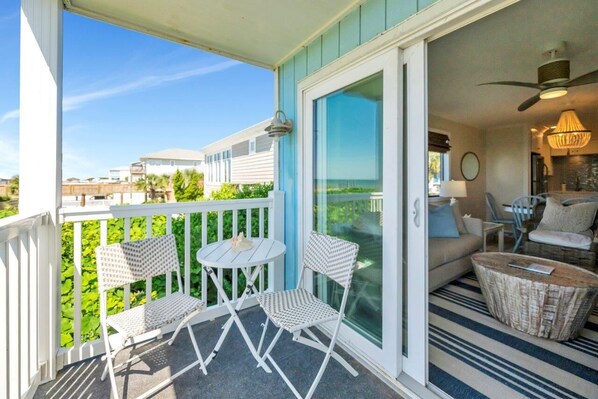 A balcony with white chairs and a table, a sliding glass door leading to a living area with a sofa, a lamp, and a ceiling fan.