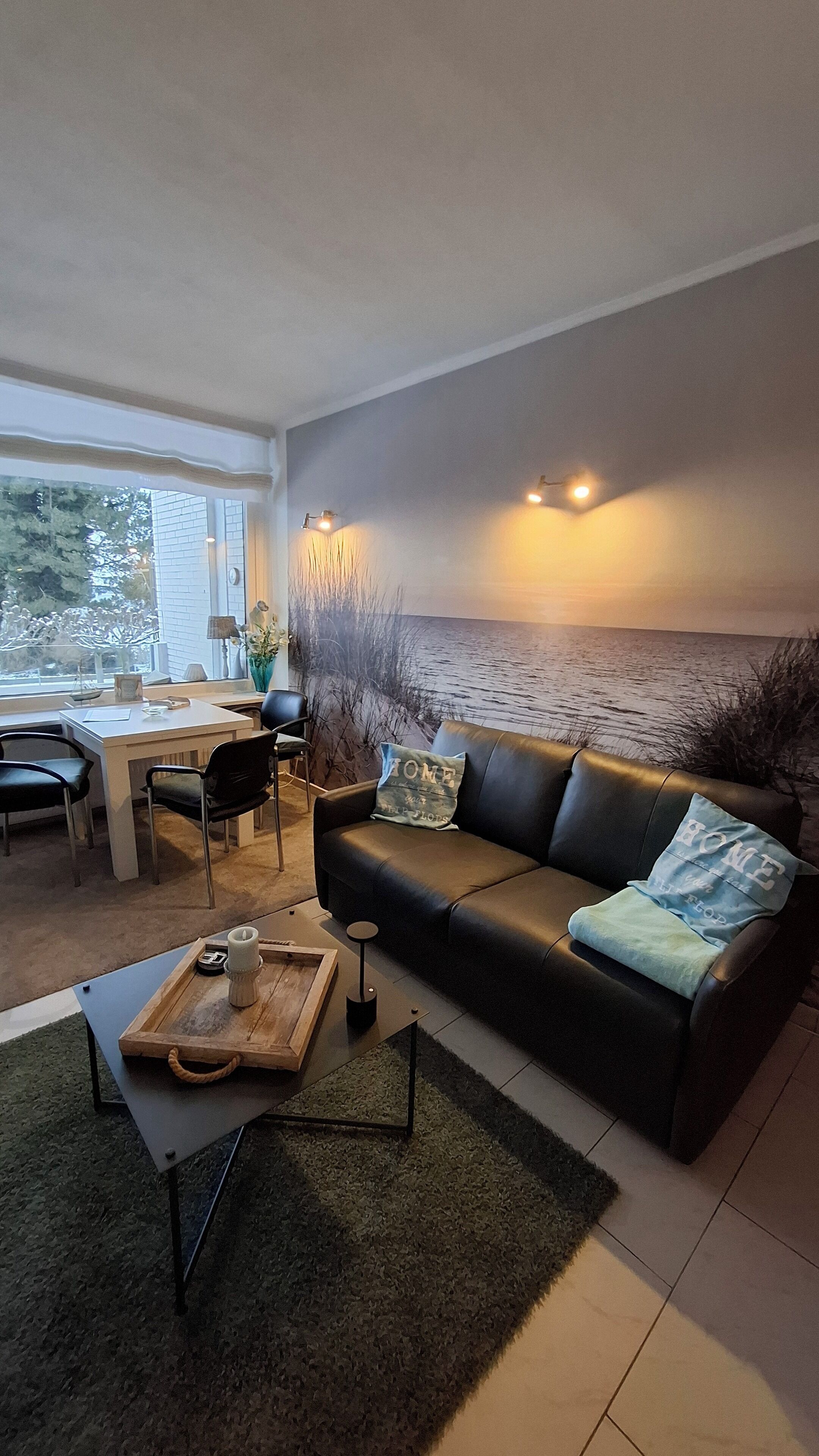 A modern living room with a black leather sofa, a wooden coffee table, and a view of the sea through a large window.