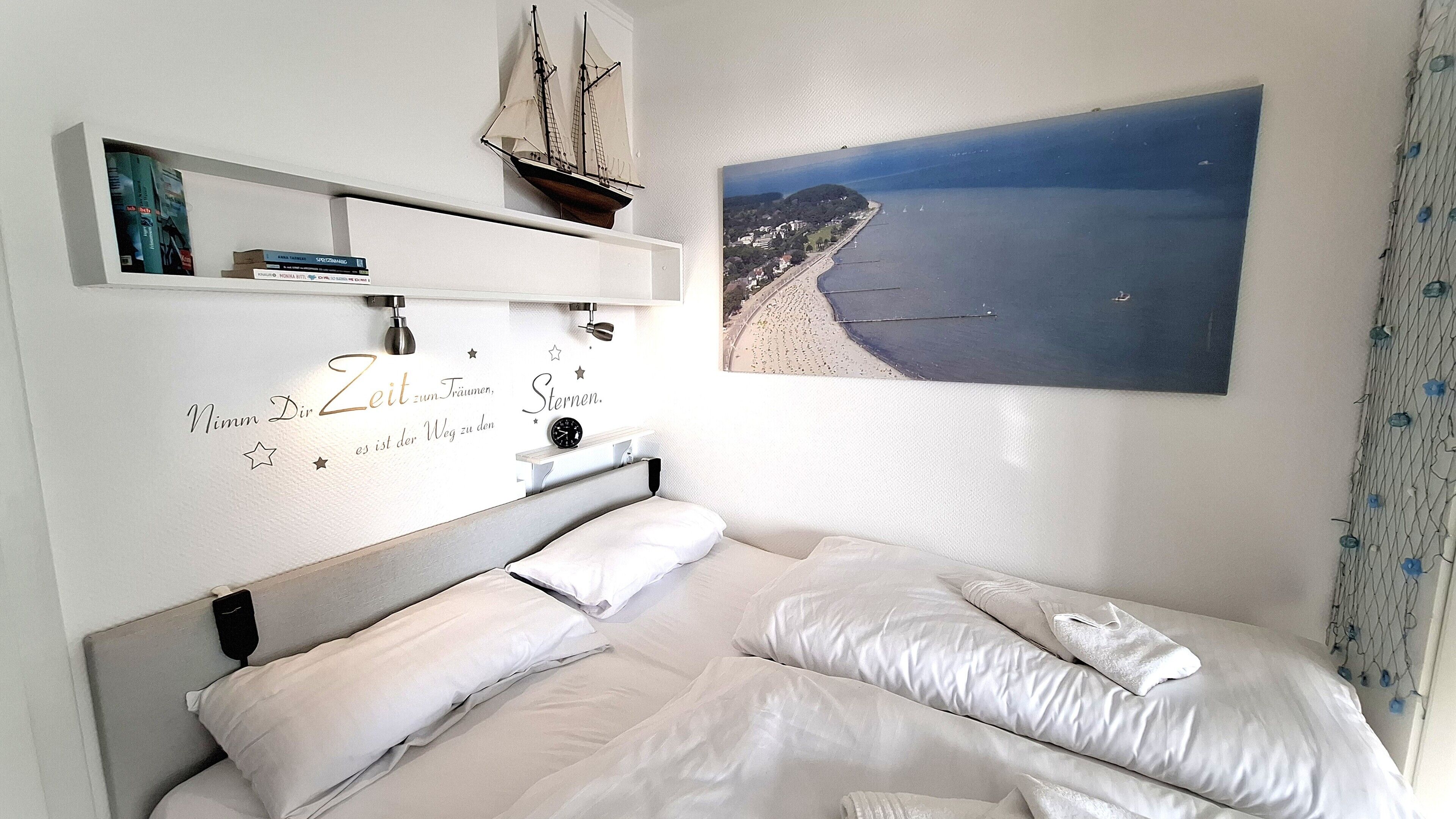 A bedroom with a bed, a wall-mounted shelf with books, a ship model, and a beach view painting.