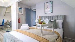 Een moderne hotelkamer met een groot bed, een nachtkastje met een fruitplateau, een ingelijst schilderij aan de muur en een blauwe deur op de achtergrond.