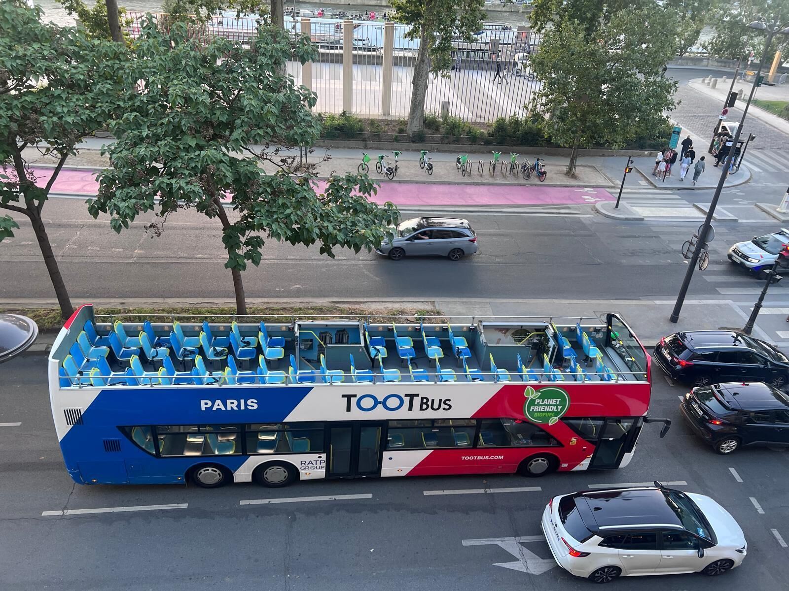 Un autobus à deux étages arborant les marques « TOOTBUS » et « PARIS », stationné à un carrefour de la ville, avec des voitures et des piétons aux alentours.