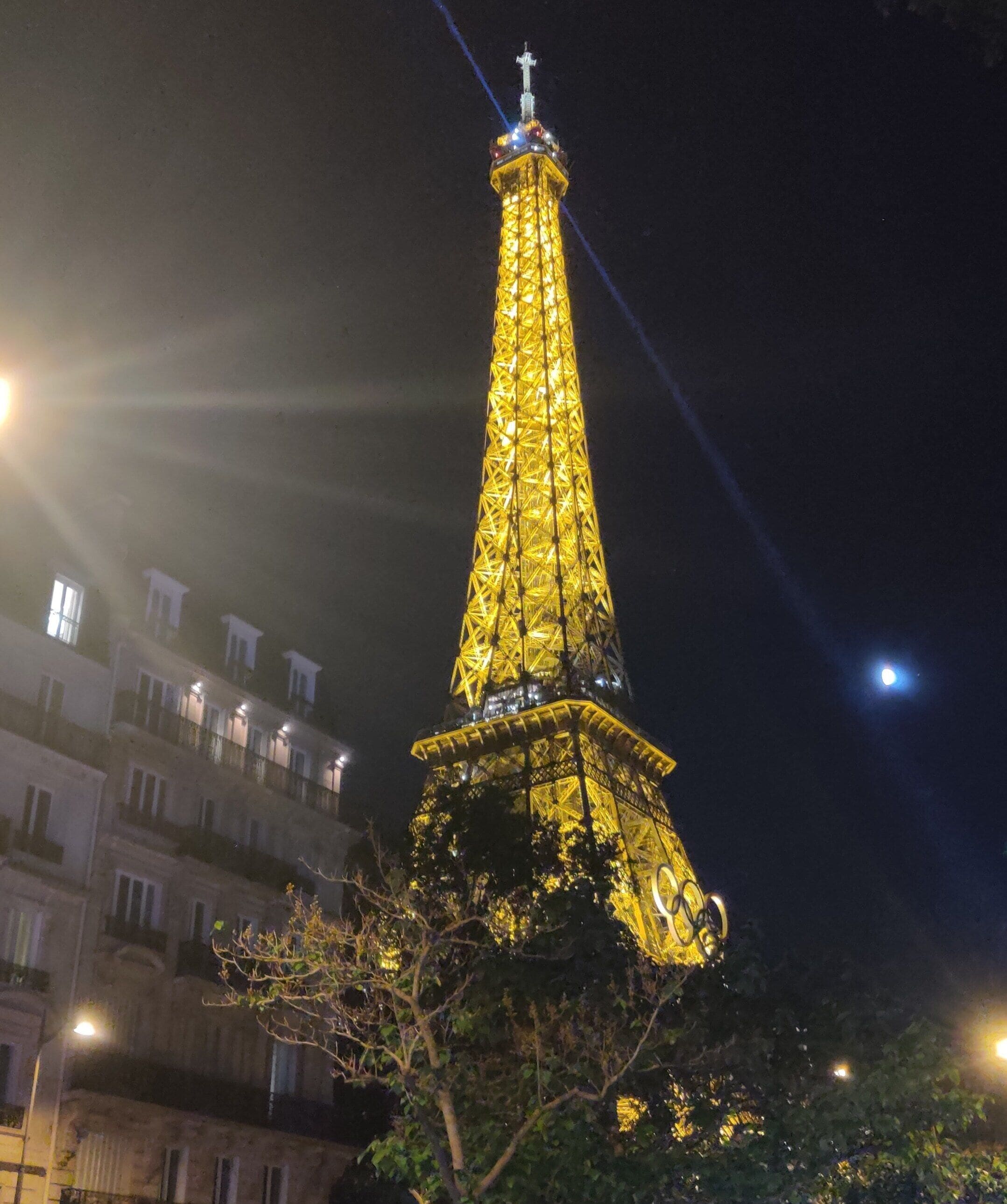 Une haute tour illuminée, surmontée d’une croix, entourée de bâtiments et d’arbres.