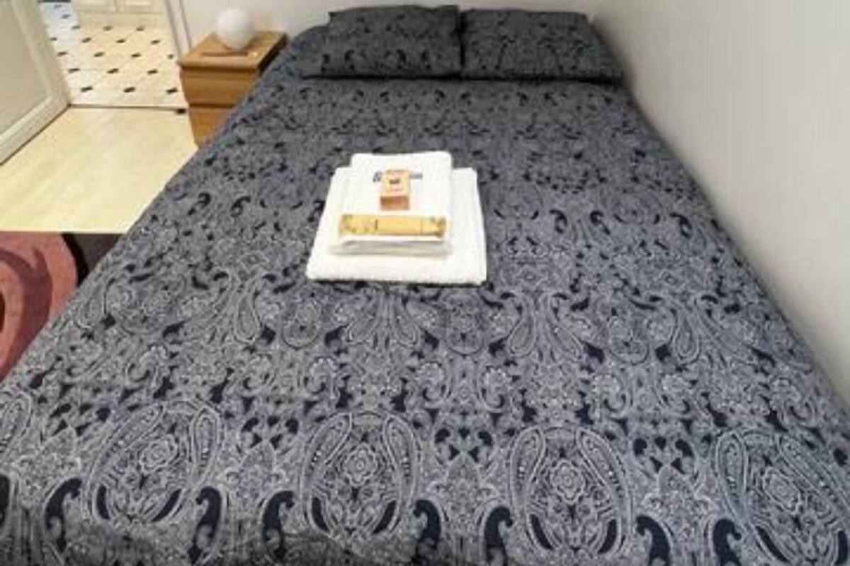 Una cama bien hecha con un edredón estampado y almohadas, una mesita de noche con una lámpara, y una puerta que da a otra habitación.