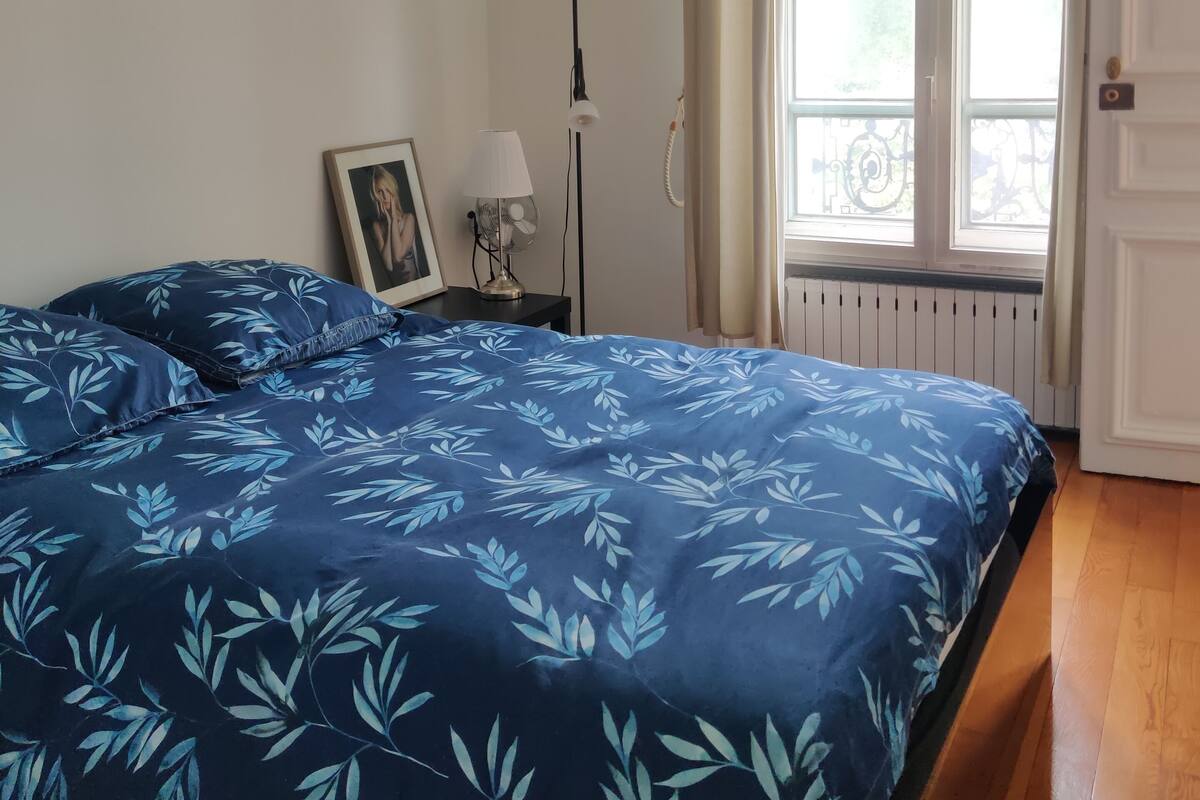 Un dormitorio con una cama grande, una lámpara de noche, un cuadro enmarcado y una ventana con cortinas.