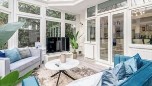 A conservatory with a white sofa, blue cushions, a round table, and a large potted plant. - Modern studio near Wembley #2 (Harrow)