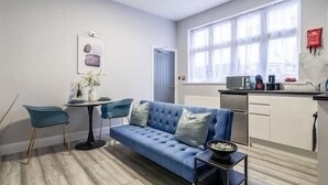 A modern living room with a blue sofa, a small round table, and a kitchen area with a microwave and coffee maker. - Modern studio near Wembley #2 (Harrow)