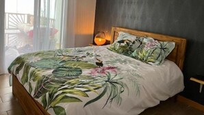 Een slaapkamer met een bed, een nachtkastje, een lamp en uitzicht op de natuur door een raam met gordijnen.
