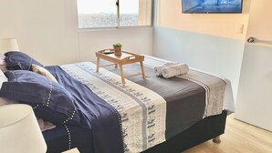 A neatly made bed with a patterned bedspread, a small wooden table with a plant, and a pair of towels. A television is mounted on the wall displaying a snowy landscape. - Private apartement with ocean view (La Perla callao)