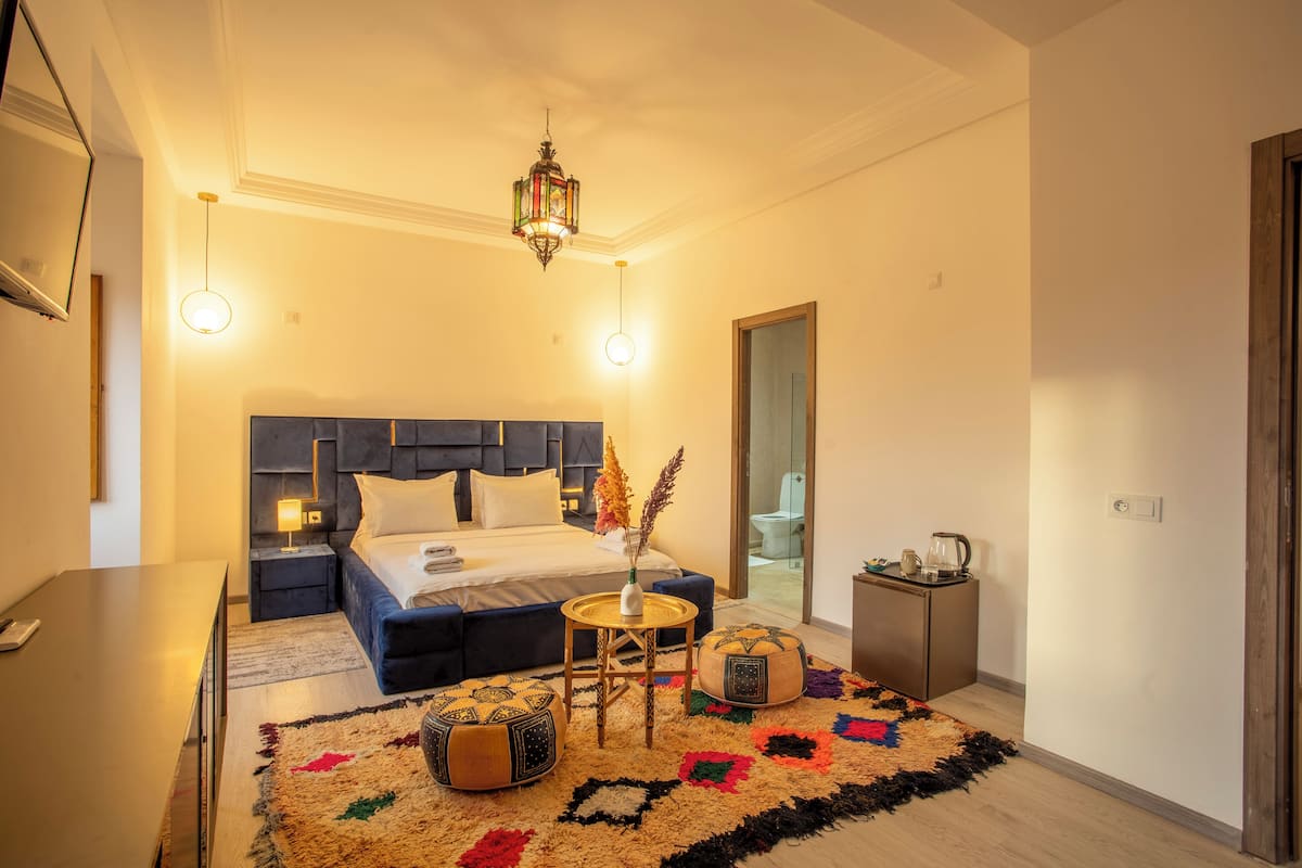 A modern hotel room with a large bed, bedside tables, a small round table with a vase, two ottomans, and a colorful rug.