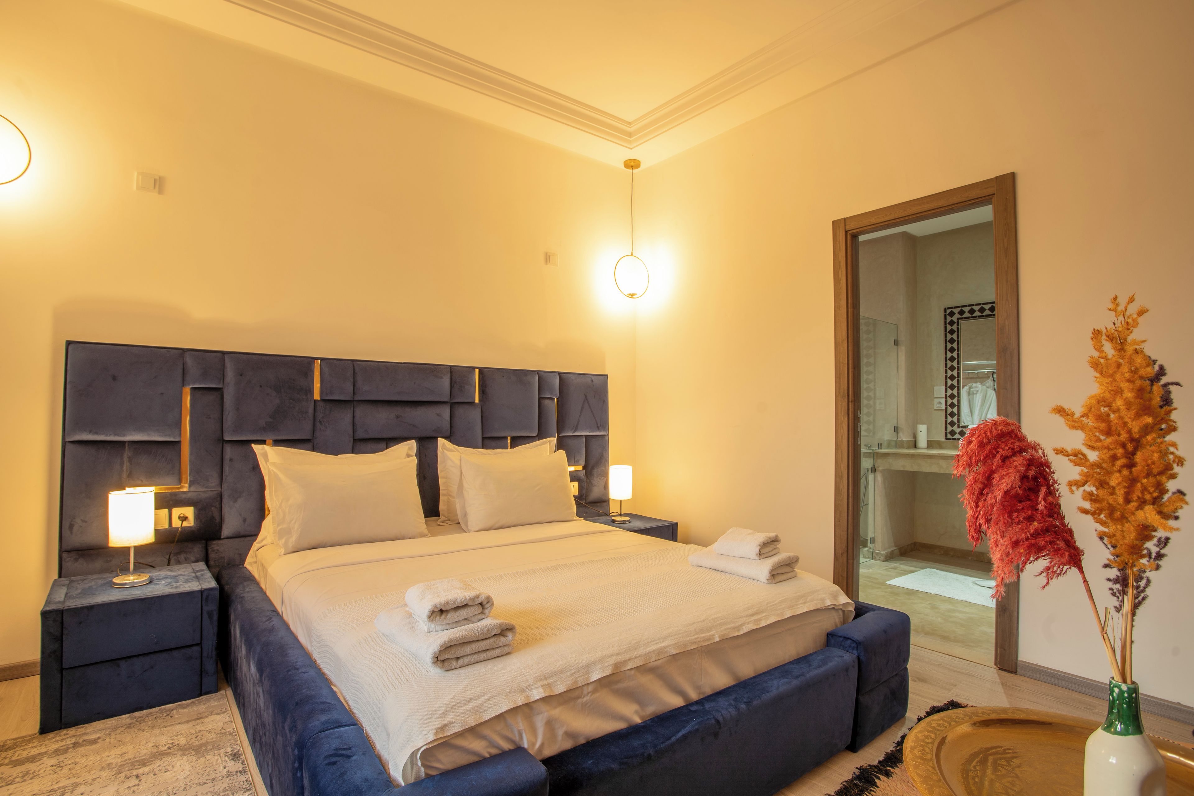 Una cama bien hecha con almohadas y toallas dobladas, una mesita de noche con lámpara y un espejo en la pared.