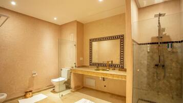 A bathroom with a beige tiled floor, a white sink, a glass shower enclosure, and a large mirror with a decorative frame.