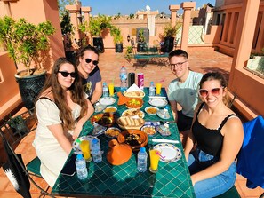 A group of people seated at an outdoor table with a variety of dishes and drinks, surrounded by potted plants and architectural features.