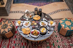 A round dining table with a variety of dishes, including bread, eggs, and fruits, set on a patterned rug. - Riad kenza (Marrakech)