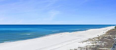 A beach with clear blue water and a sandy shore. There are steps leading down to the beach, and a few people are visible in the distance.