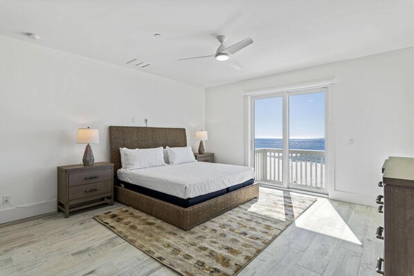 A bedroom with a large bed, two bedside tables with lamps, a ceiling fan, and a view of the ocean through a sliding glass door.