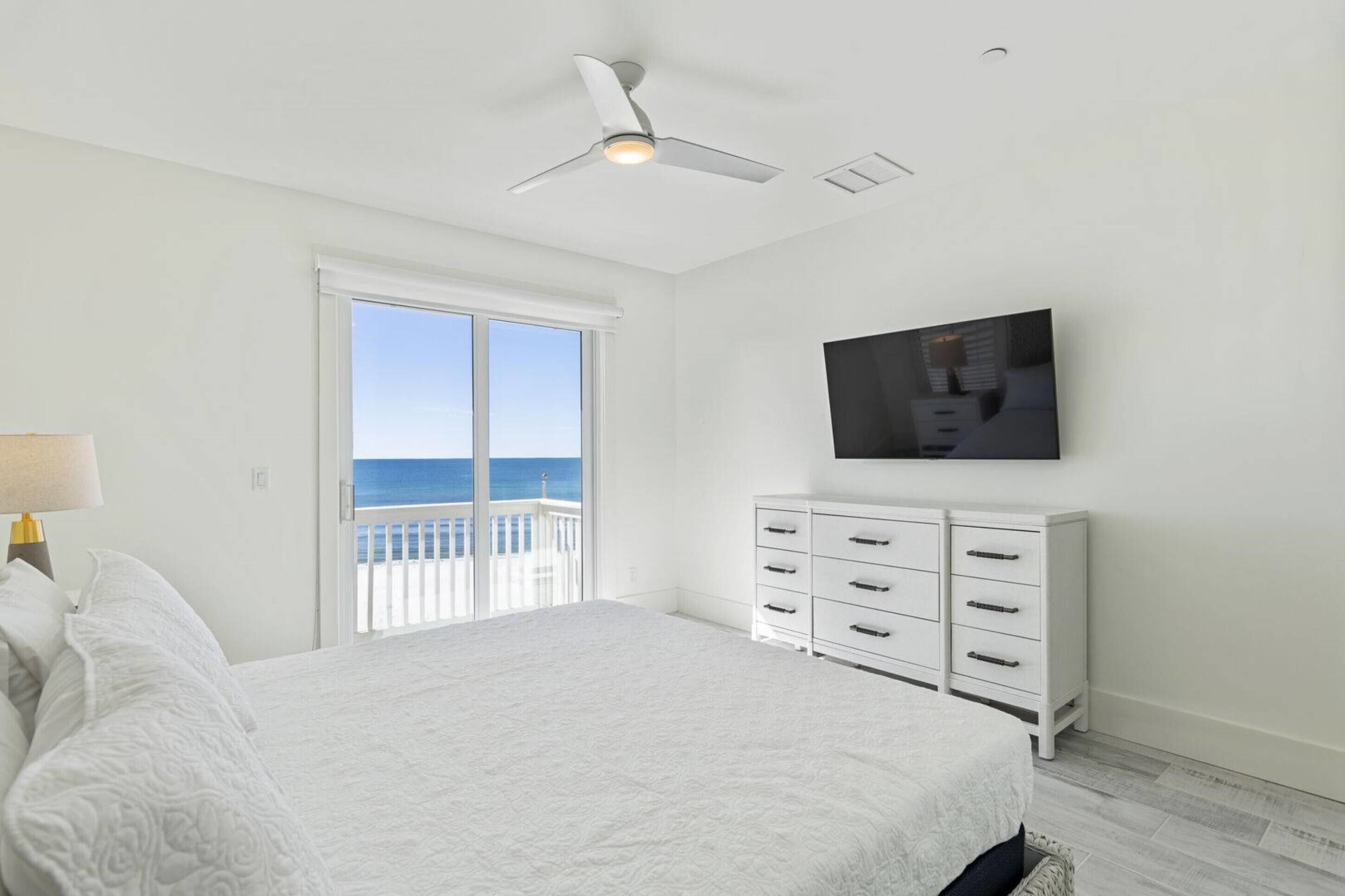 A bedroom with a bed, a ceiling fan, a TV mounted on the wall, a white dresser, and a balcony with a view of the ocean.
