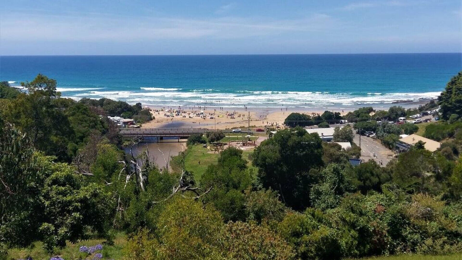 A coastal view with a beach, a bridge, and dense greenery.