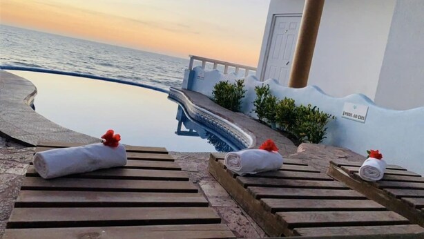 Ein Poolbereich mit Holzliegen, einem Pool und Blick auf das Meer bei Sonnenuntergang.