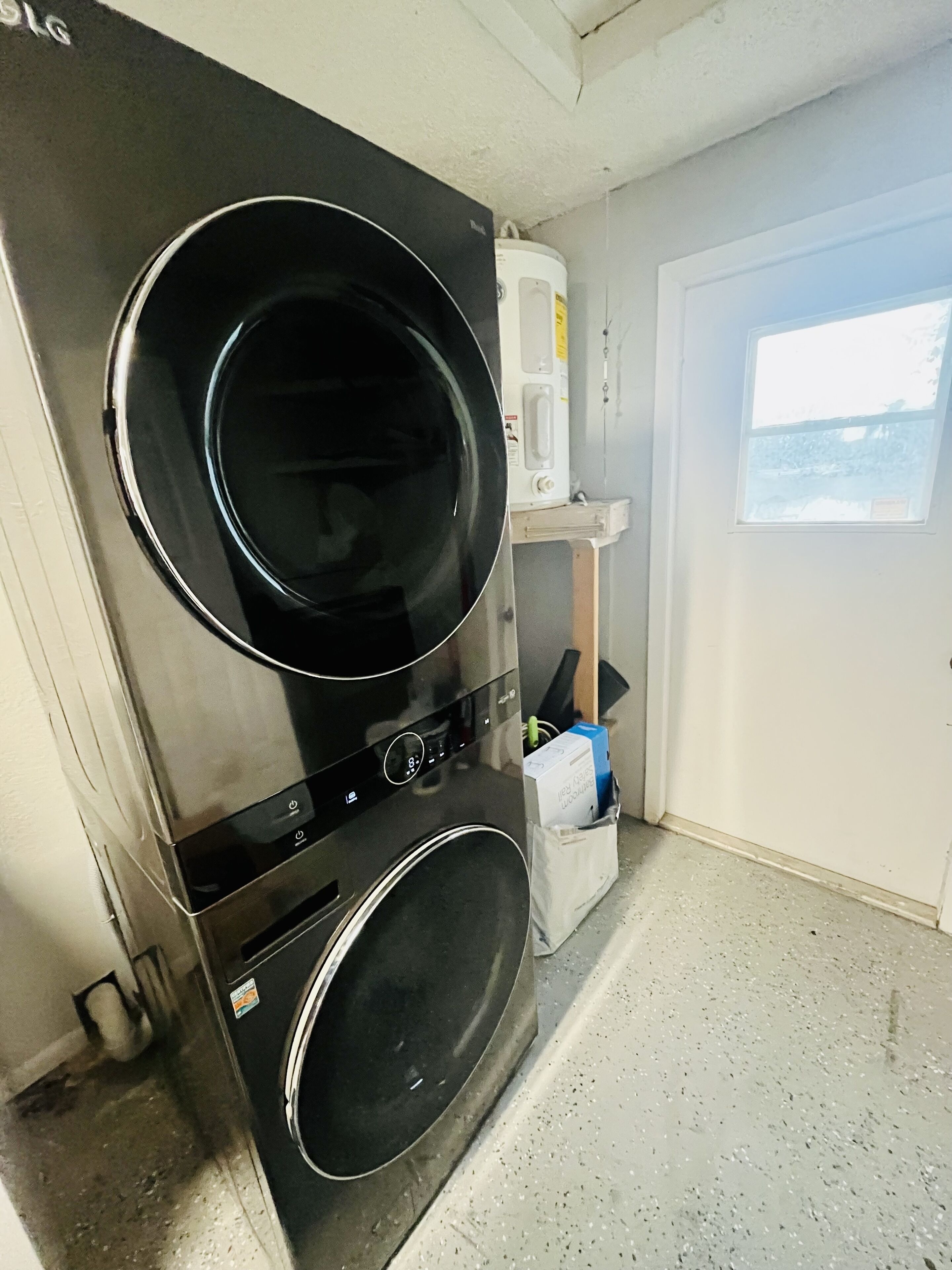 Un área de lavandería compacta con lavadora y secadora de carga frontal, un estante pequeño con calentador de agua y una puerta que da a un balcón.