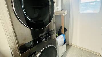 Un área de lavandería compacta con lavadora y secadora de carga frontal, un estante pequeño con calentador de agua y una puerta que da a un balcón.