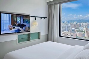 A hotel room with a bed, a TV mounted on the wall, a window with a city view, and a hanging lamp.