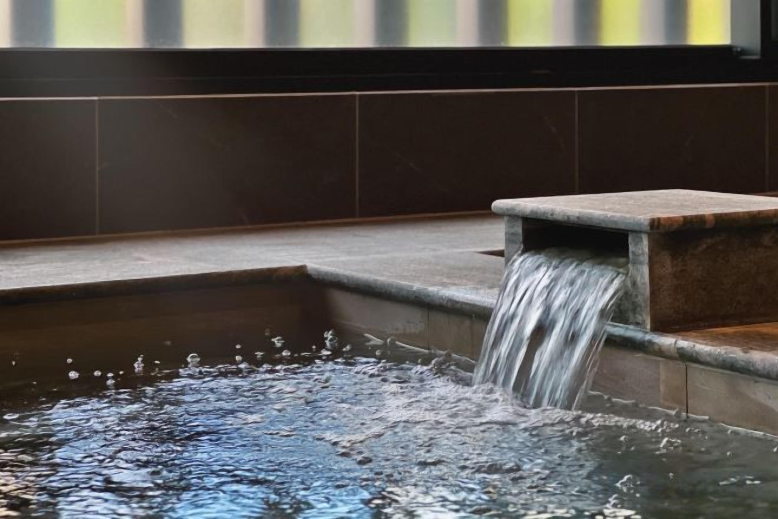 Ein Wasserspiel mit einem kaskadenartigen Wasserfall in ein rechteckiges Becken, vor einer mit Fliesen verkleideten Wand.