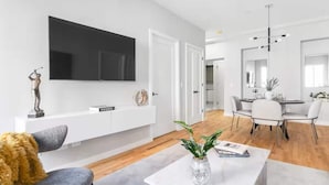 Ein modernes Wohnzimmer mit einem Flachbildfernseher, ein Essbereich mit einem weißen Tisch und Stühlen, und ein Gang, der zur Küche führt.