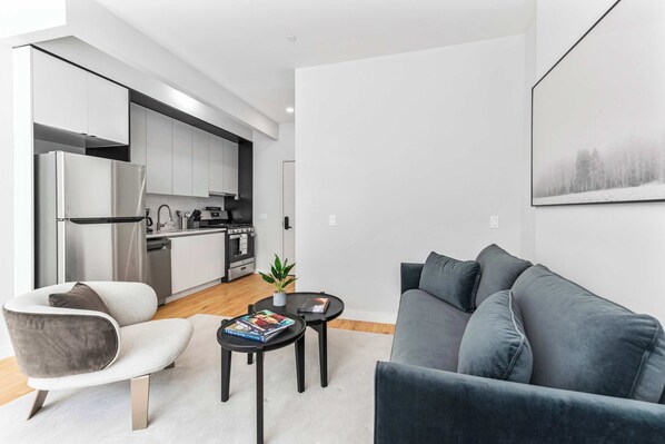 A modern living space with a grey sofa, a white armchair, a small round table with a book, and a kitchen area with white cabinets and stainless steel appliances.