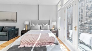 A modern bedroom with a large bed, grey and white decor, a grey sofa, and a city view through large windows. - Luxe Studio with Charming Juliet Balcony (New York)