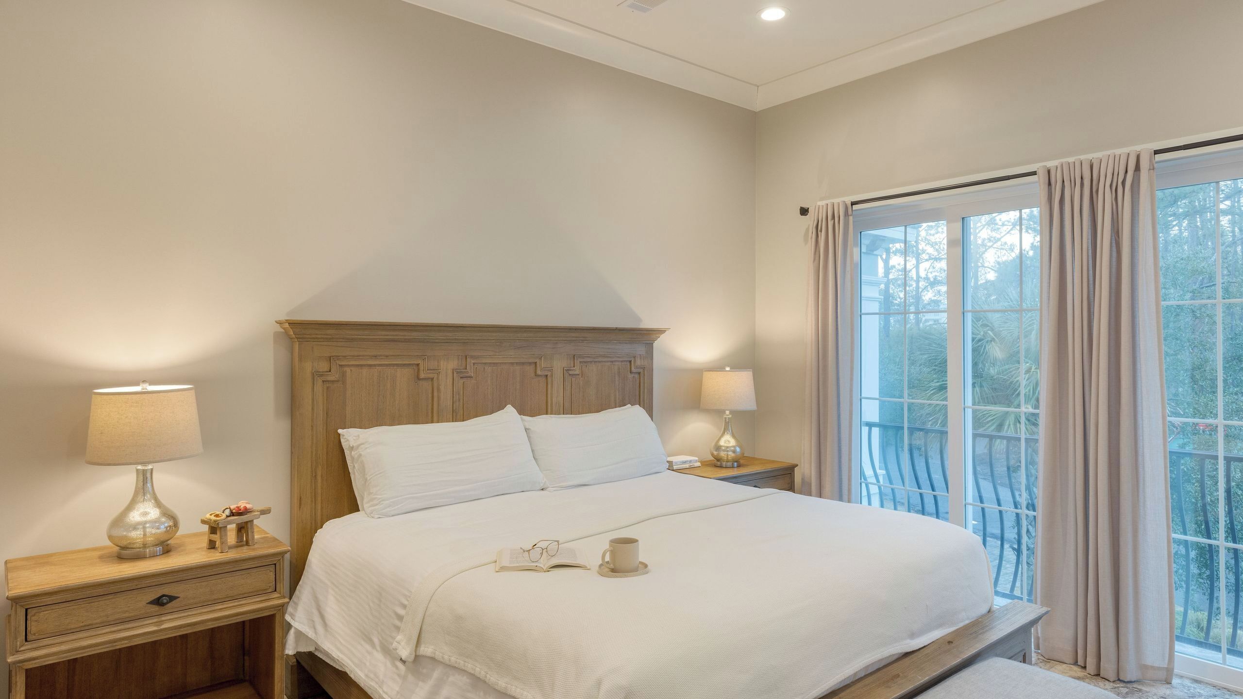 A bedroom with a wooden headboard, two bedside tables with lamps, a bed with white bedding, and a view of greenery through the window.