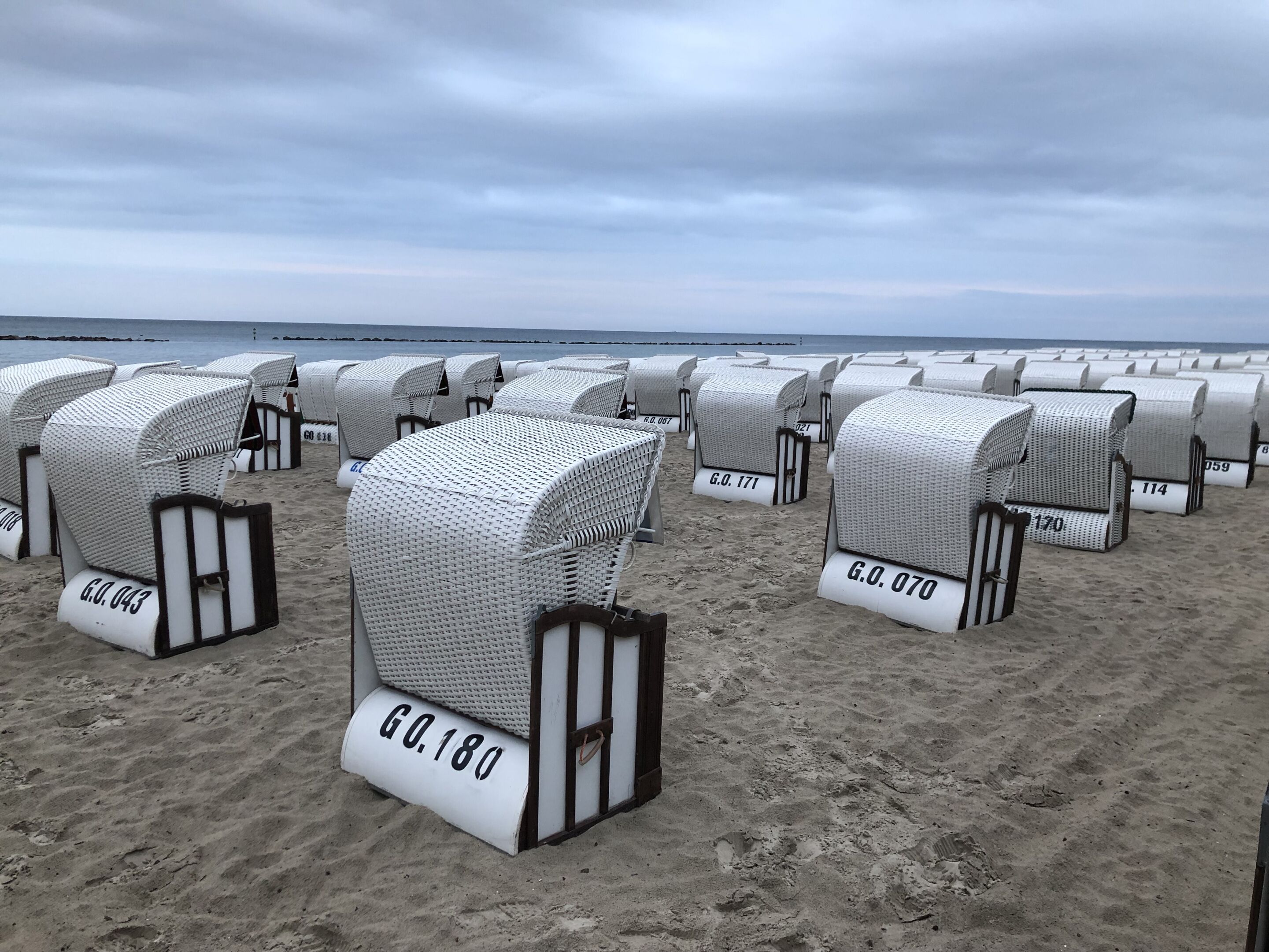 Filas de sillas de mimbre junto a la playa, con números en la base, dispuestas sobre arena, con el mar al fondo.