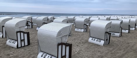 Filas de sillas de mimbre junto a la playa, con números en la base, dispuestas sobre arena, con el mar al fondo.